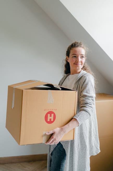 A person inside a home is in the process of packing or preparing for a move, placing a cardboard box on top of other sealed boxes. The individual is wearing a white T-shirt and light-colored trousers,