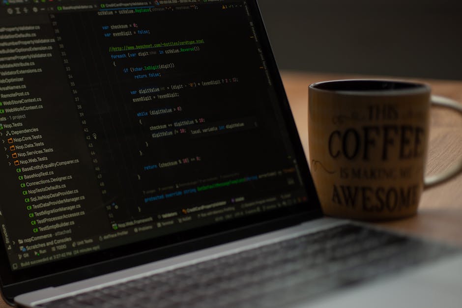 A close-up view of a laptop computer displaying lines of code on its screen, placed on a wooden desk. To the right of the laptop is a coffee mug with a printed message that reads 'This coffee is makin