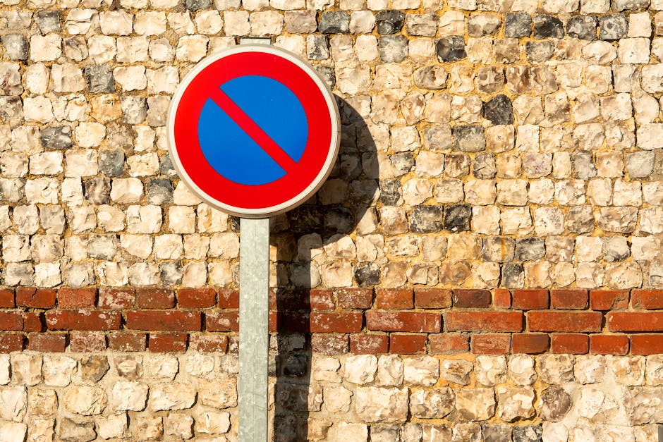 A circular blue and red 'no parking' sign mounted on a metal pole is positioned in front of a textured stone wall with a horizontal red brick band running across the middle. The sign casts a shadow on